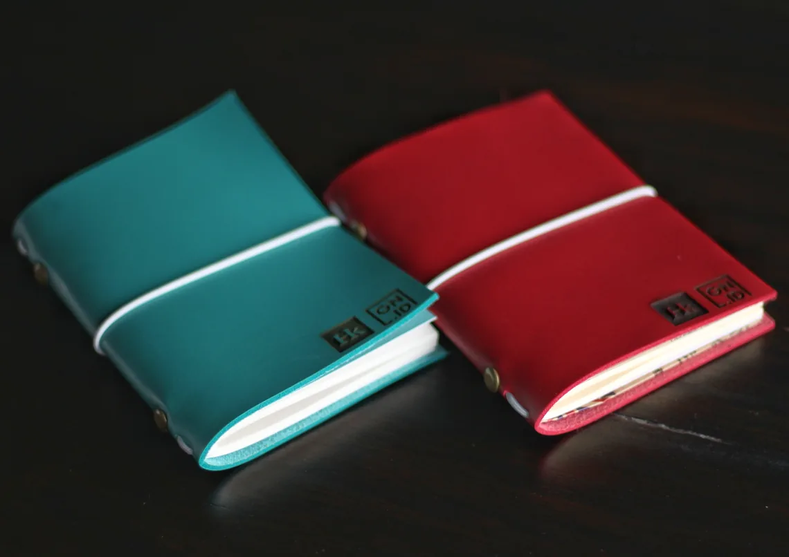 Two leather travel journal covers standing on a dark surface, one teal and one red, each closed with a white elastic band and carrying a small dark label with two logos