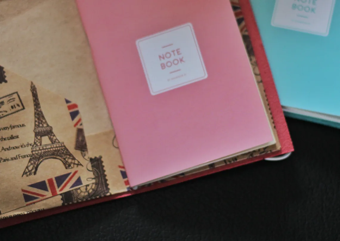 A red leather cover opened flat to show three stacked paper inserts: a kraft insert printed with Eiffel Tower and Union Jack travel stamps, a pink insert labelled Note Book, and a pale blue insert behind